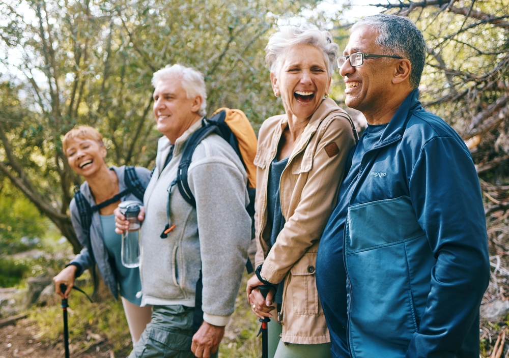 Four older adults, two women and two men, are smiling and laughing together while hiking outdoors in a forested area, dressed in casual clothing and carrying backpacks—enjoying how the most effective medication for osteoarthritis supports their active lifestyle.