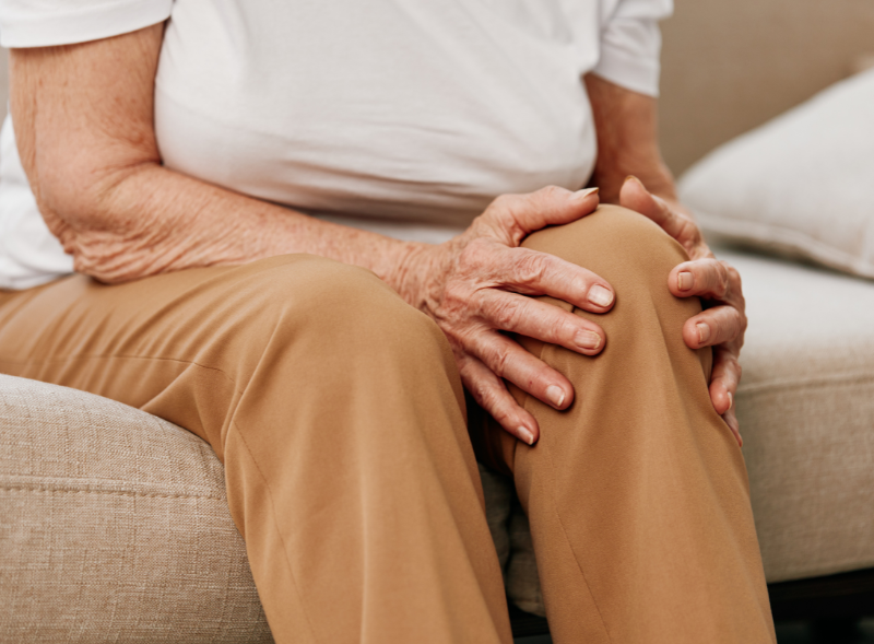 An older adult with joint pain in the cold weather, wearing a white shirt and beige pants sits on a couch, holding their knee with both hands, suggesting discomfort or knee pain.