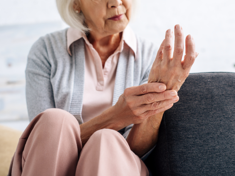 An elderly woman with joint pain during cold weather, sitting on a couch holds and massages her painful wrist with one hand, suggesting discomfort or arthritis. She wears a light pink blouse, gray cardigan, and beige pants.