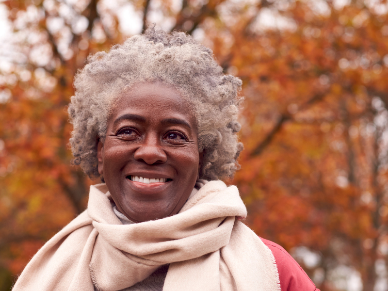 An older woman with joint pain in the cold weather, with gray curly hair smiles warmly outdoors, wearing a light beige scarf and jacket. The background is filled with autumn trees in shades of orange and brown.