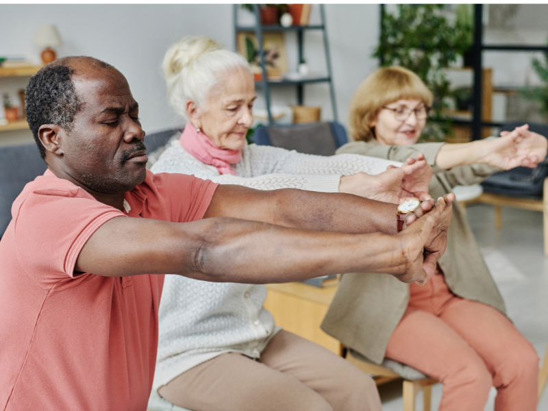 Three older adults preventing joint pain during cold weather, sitting indoors, stretching their arms forward in unison. They appear focused and relaxed, participating in a group exercise or wellness activity in a bright, comfortable setting.