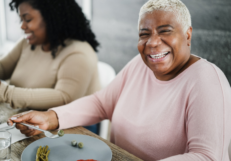 Two women sit at a table eating together. The woman in the foreground has joint pain in the cold weather, wearing a light pink top, smiles brightly at the camera while holding a fork with green olives. The background woman is blurred and also eating.