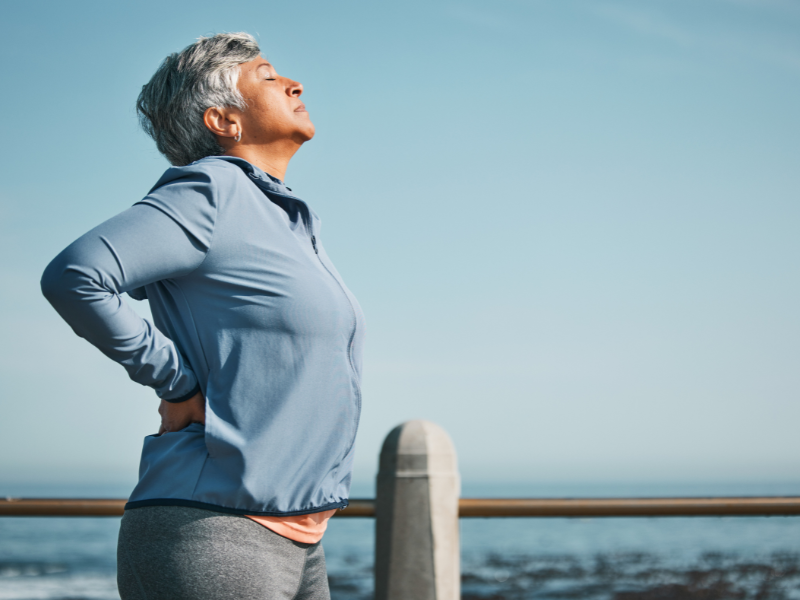 Osteoarthritis in women: Older woman with short gray hair stands outdoors by the ocean, eyes closed, hands on lower back, appearing to stretch or relieve back pain, wearing a light blue jacket and exercising clothes under a clear sky.