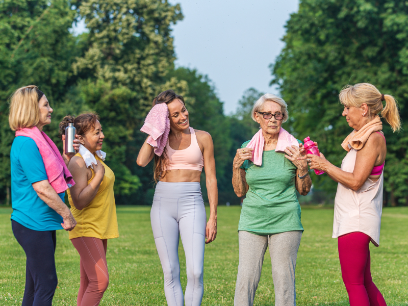 Menopause and joint pain. Five women of varying ages stand together outdoors on grass, smiling and talking after exercising. They hold water bottles and towels, dressed in activewear, with trees and a blue sky in the background.
