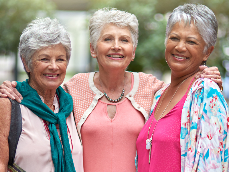 Why are females more prone to osteoarthritis? : Three smiling older women stand close together outdoors, arms around each other, wearing colorful tops and light jackets. The background is softly blurred with greenery.